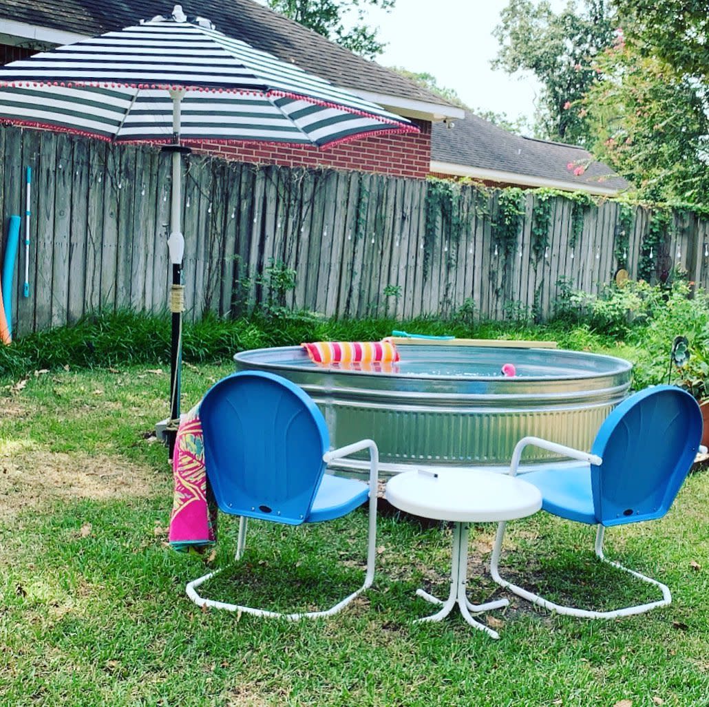 Relaxing backyard setup with a galvanized stock tank pool, striped umbrella, blue metal chairs, and a small white table on the grass