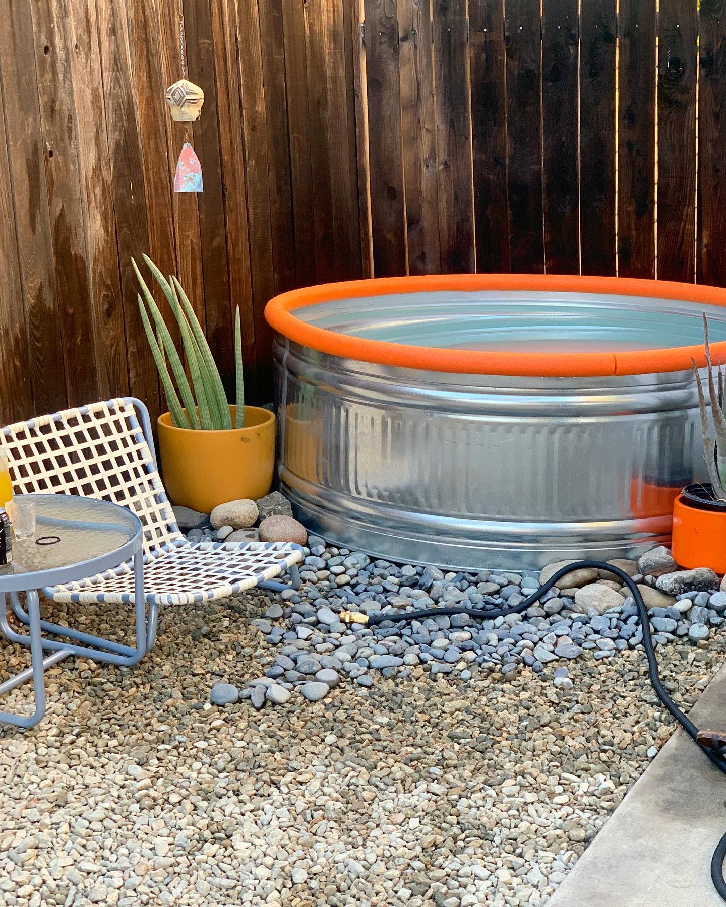 A small backyard features a round metal tub pool, a white woven chair, a gray chair, potted plants, and gravel ground covering