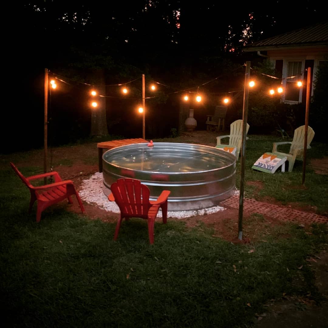 A round metal pool in a backyard with string lights above, surrounded by three colorful chairs on grass at dusk