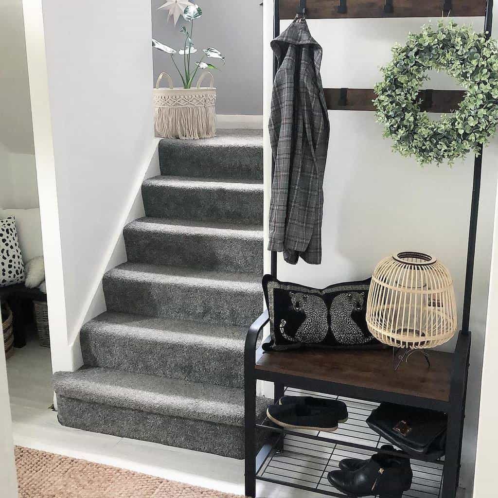 A modern entryway with gray carpeted stairs, a coat rack with a wreath and plaid jacket, a shelf with shoes, and decorative plants