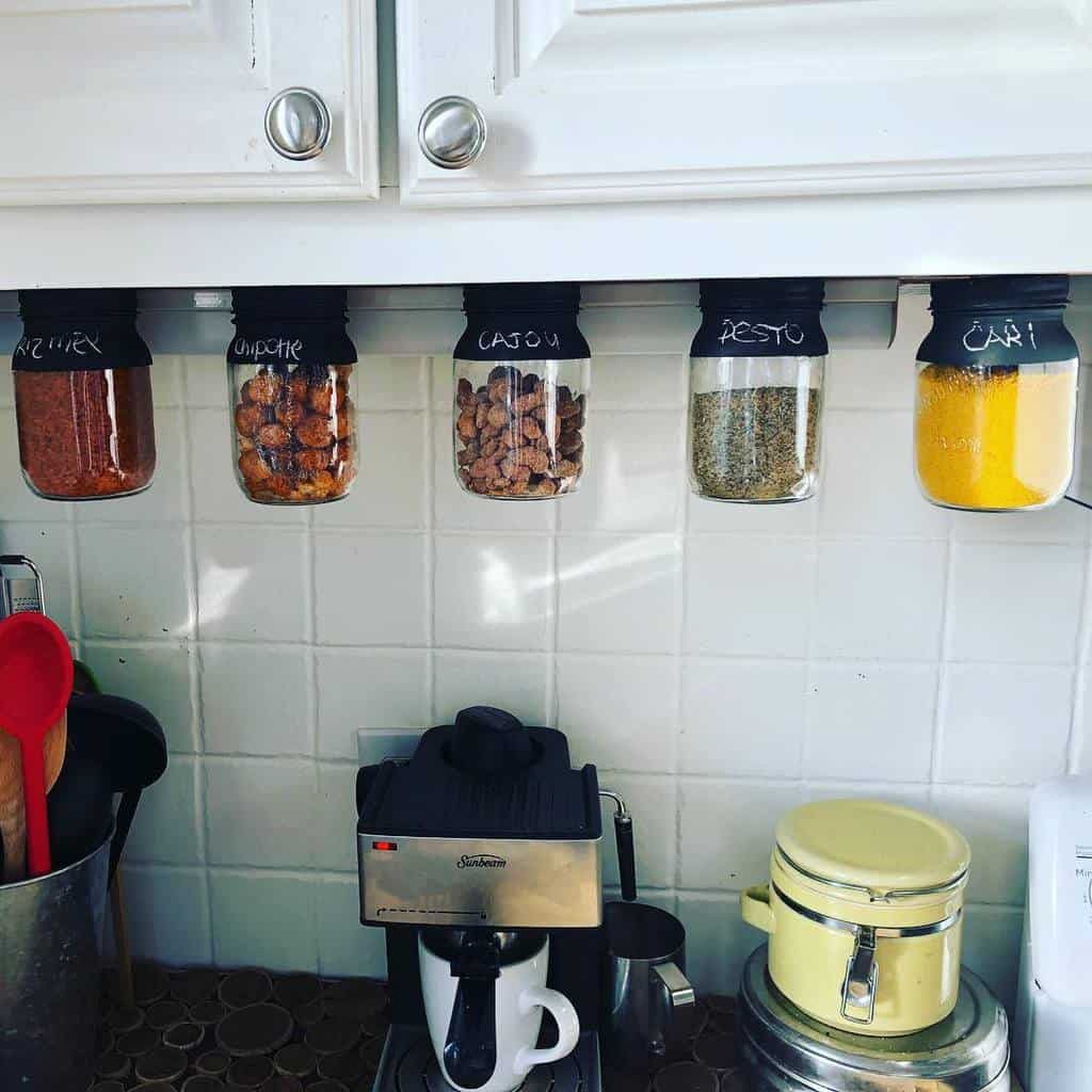 Jars of spices and nuts hanging under a kitchen cabinet, over a coffee maker and kitchen countertop appliances