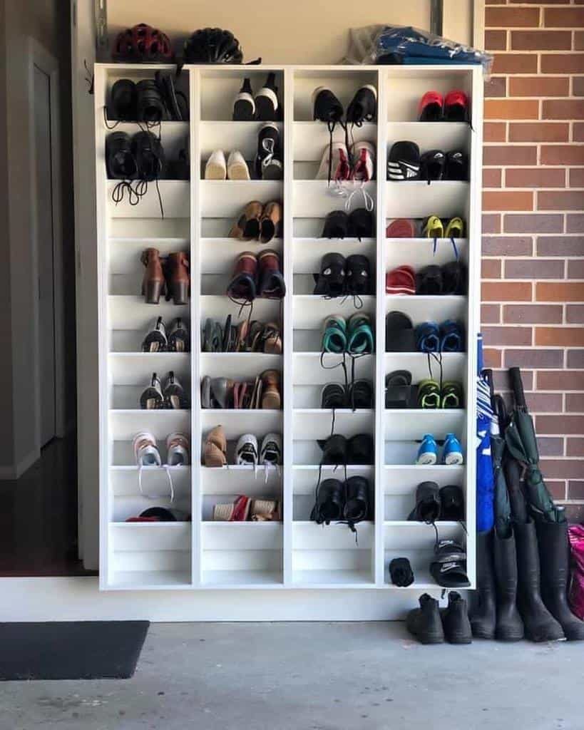 A white shoe rack holding various pairs of shoes is against a brick wall, with boots and umbrellas neatly arranged beside it