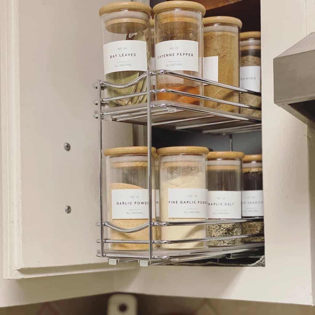 Glass jars with wooden lids containing spices like bay leaves and cayenne pepper are organized on metal shelves inside a white cabinet