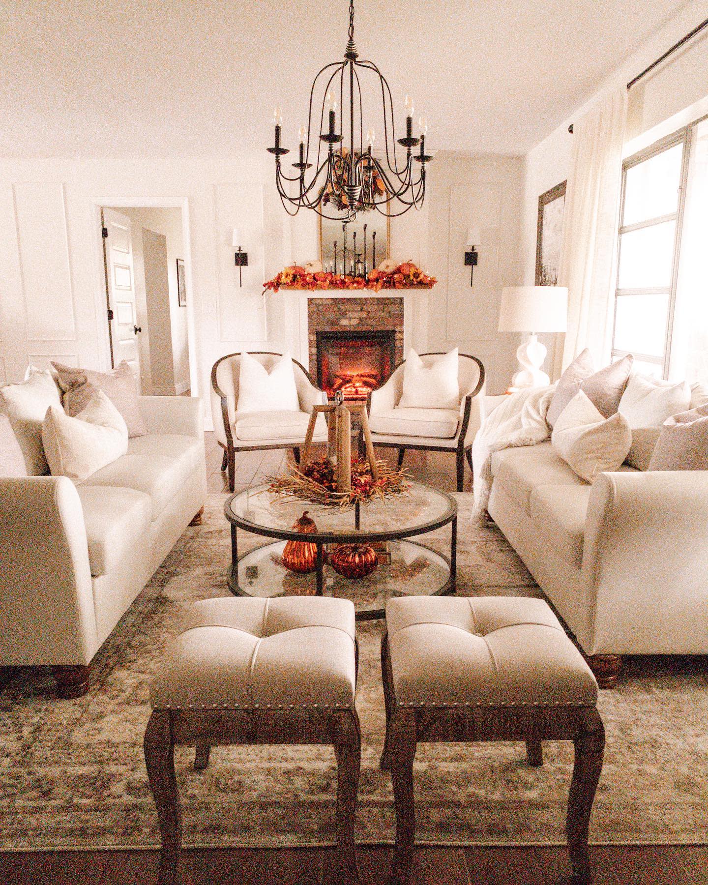 Cozy living room with white sofas, a glass coffee table, autumn-themed decor, a chandelier, and a fireplace adorned with orange garlands