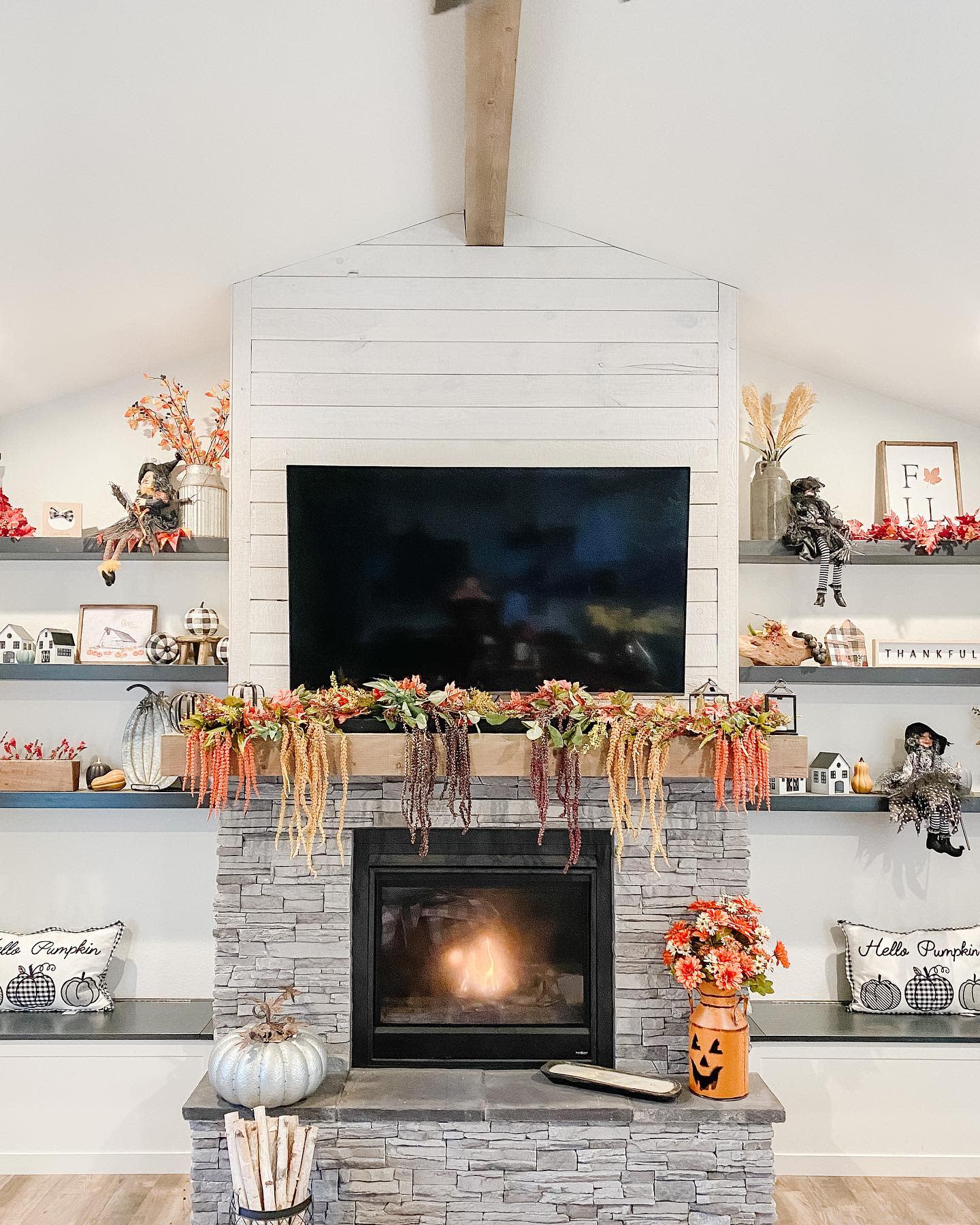 Cozy fireplace with fall decorations, including pumpkins, autumn leaves, and a garland; TV above, and shelves with festive decor