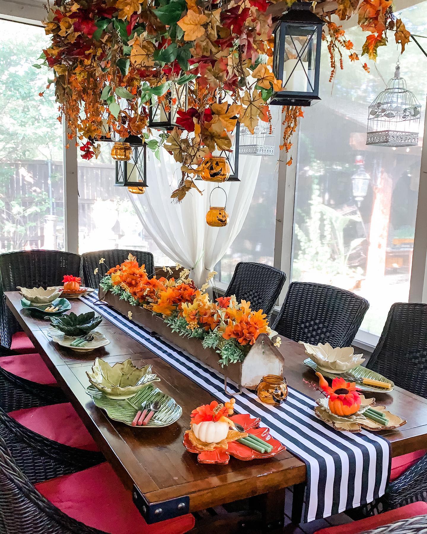 Festive fall table setting with leaf-shaped plates, pumpkins, striped runner, and autumn leaves hanging above lanterns and nature in background