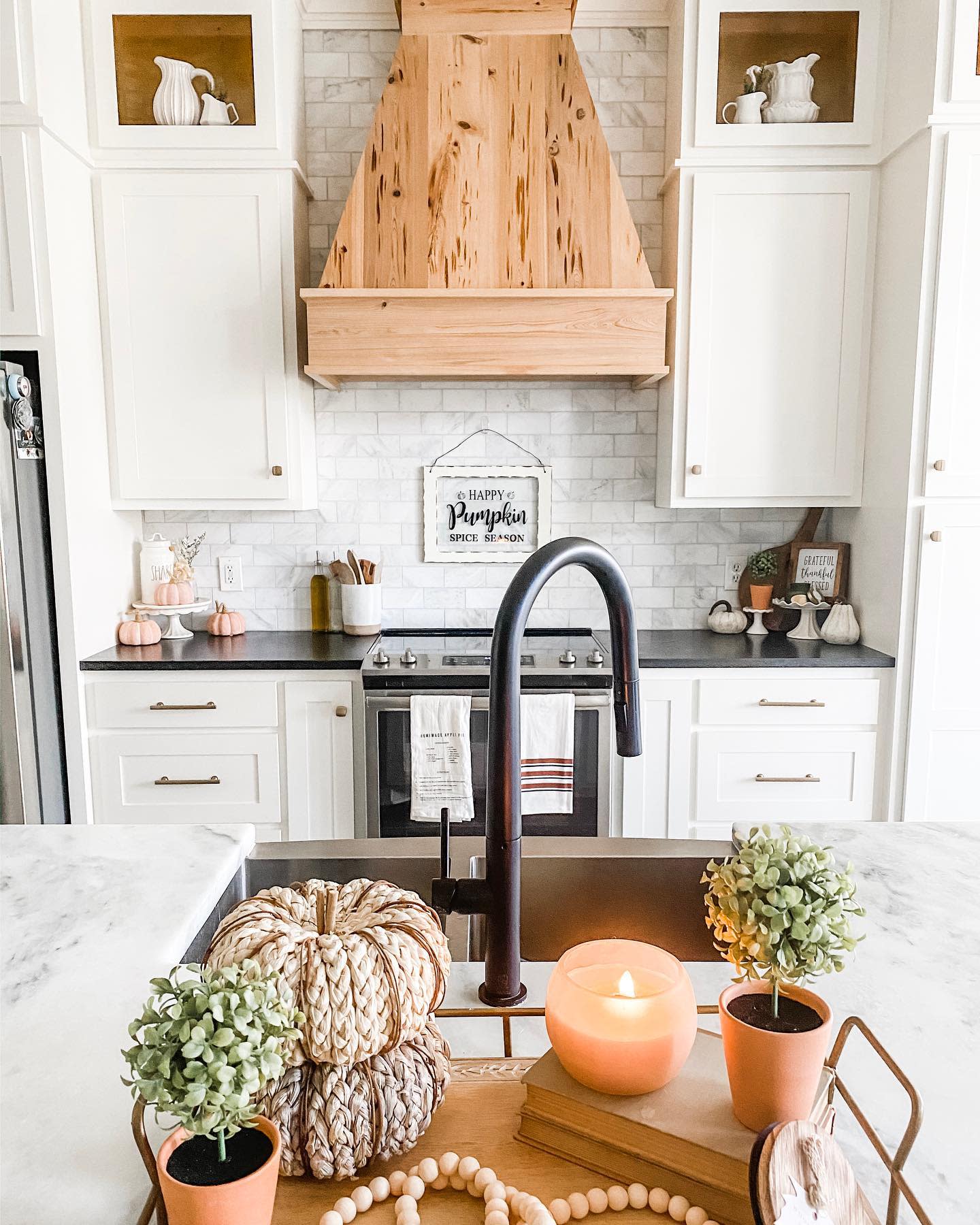 A kitchen with fall decor: knitted pumpkins, candles, and greenery on a marble island with a wooden hood above a stove with white cabinets