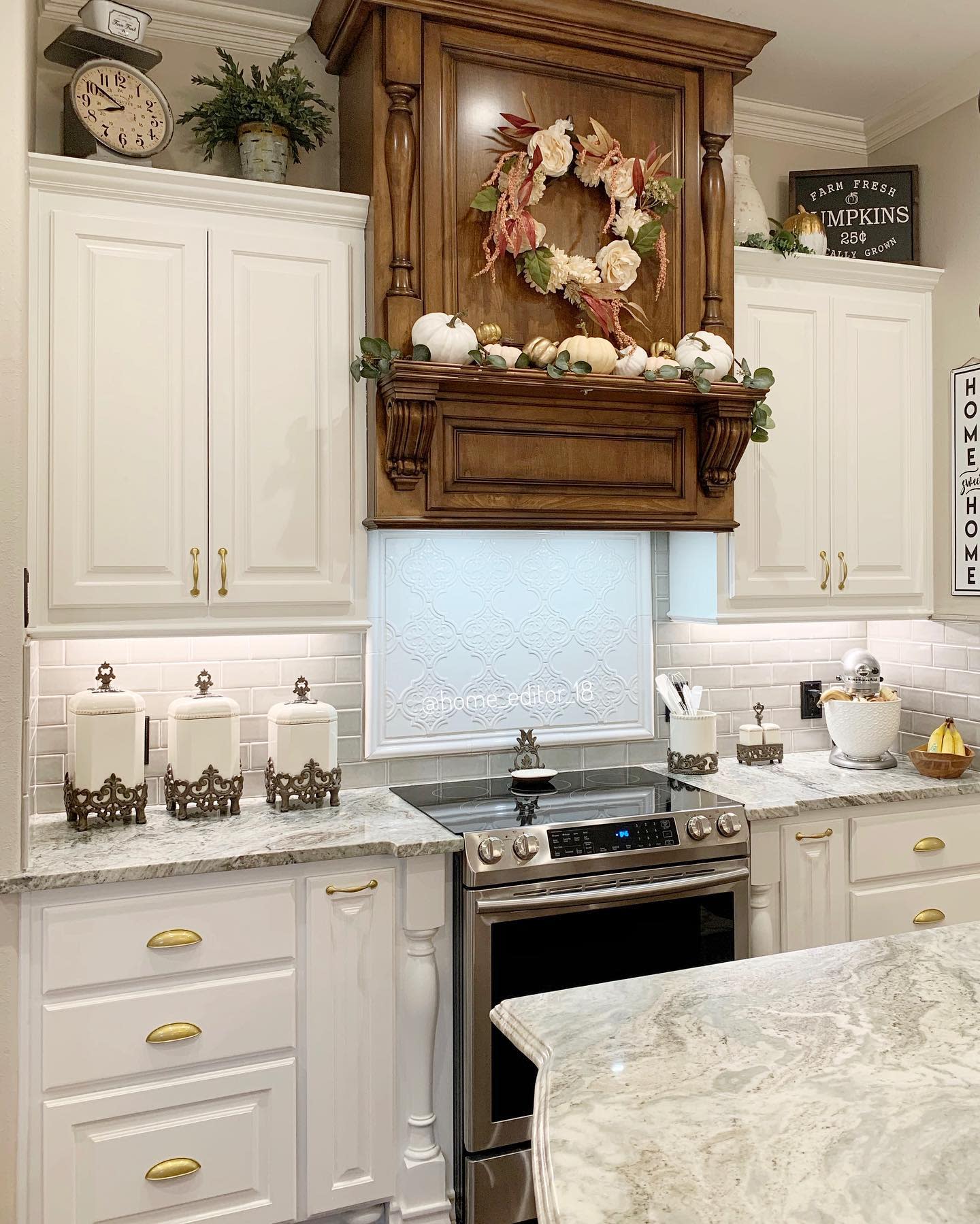 A kitchen with white cabinets, a decorative wood hood, fall wreath, pumpkins, and jars on the counter; marble countertops and stove visible