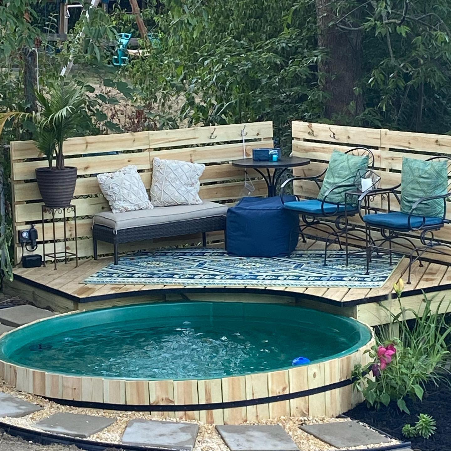 Wooden deck with a small round pool, patio furniture, cushions, potted plant, and a patterned rug surrounded by greenery