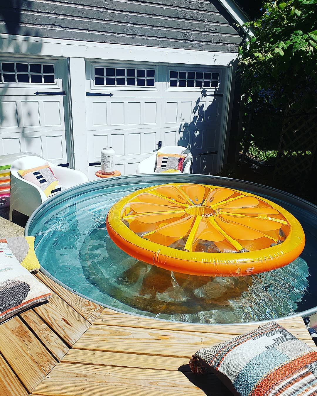 Above-ground pool with orange slice float, surrounded by patio furniture and pillows, near a garage with white doors