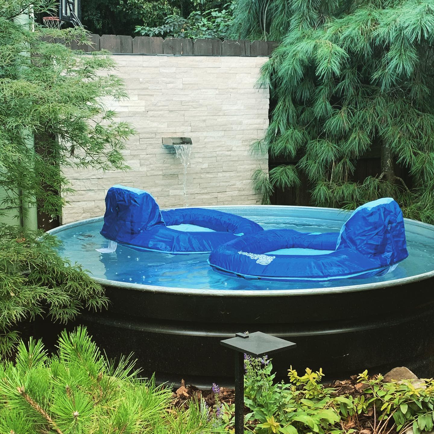 Small pool with two blue lounge chairs, surrounded by lush greenery and a stone wall with a waterfall feature