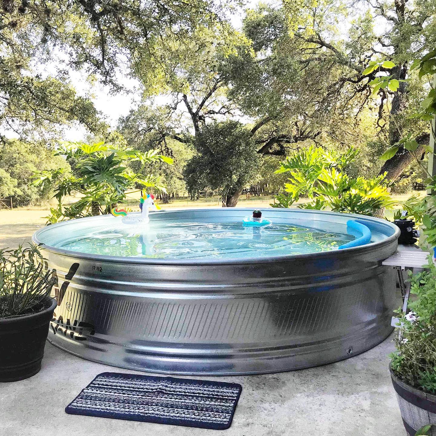 Stock tank pool surrounded by lush green plants and trees in an outdoor setting, with a striped mat on the concrete floor