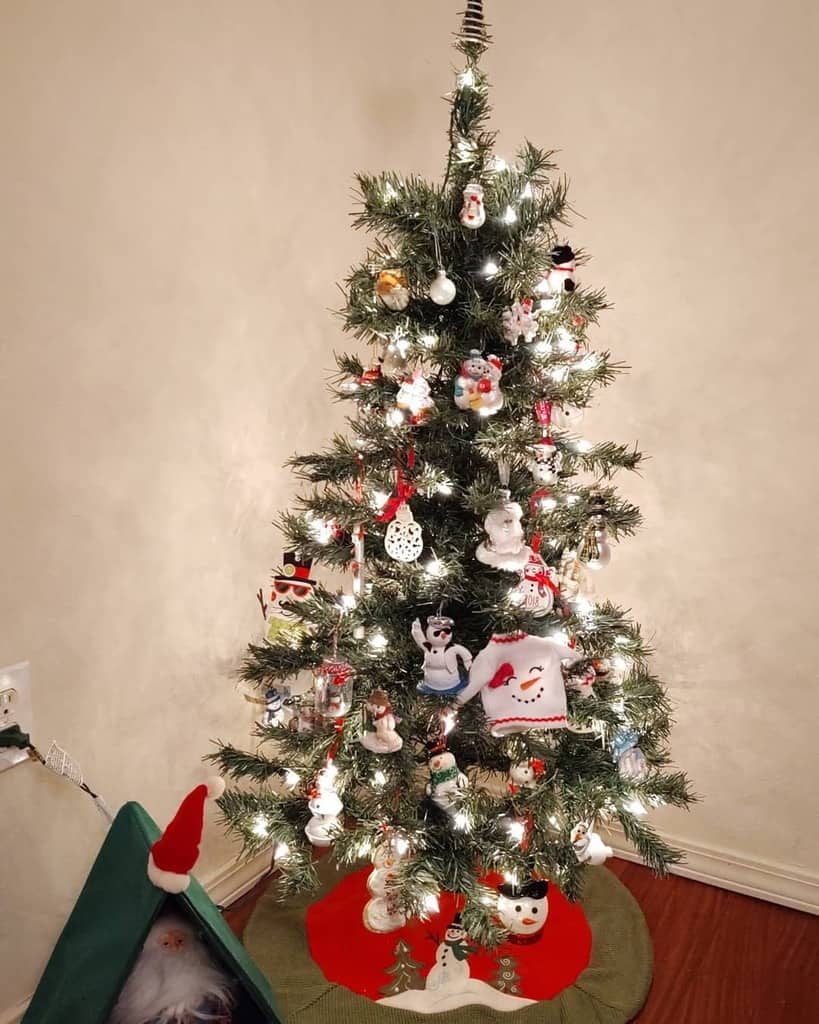 A Christmas tree adorned with lights, snowmen, and festive ornaments stands on a green and red tree skirt in a cozy room