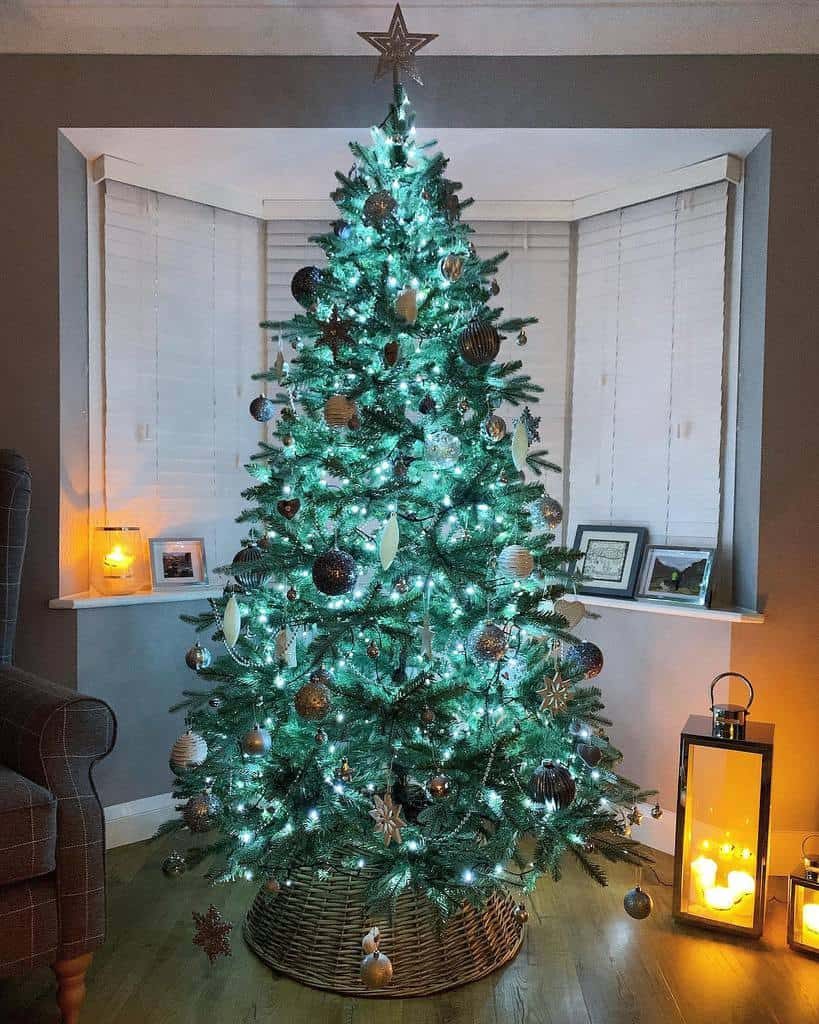 A Christmas tree with white lights and various ornaments stands by a window, with a cozy chair and lantern nearby