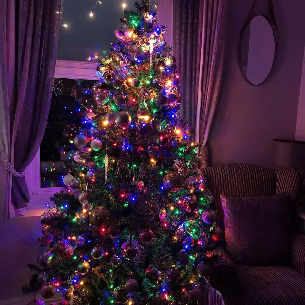 A Christmas tree with white lights and various ornaments stands by a window, with a cozy chair and lantern nearby