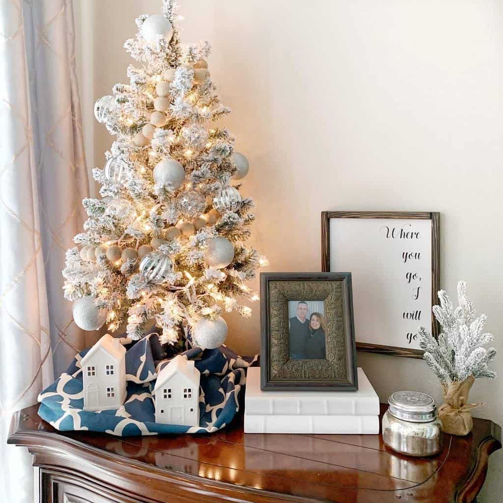 A small, decorated Christmas tree on a wooden table with framed photos, white ceramic houses, and a quote print