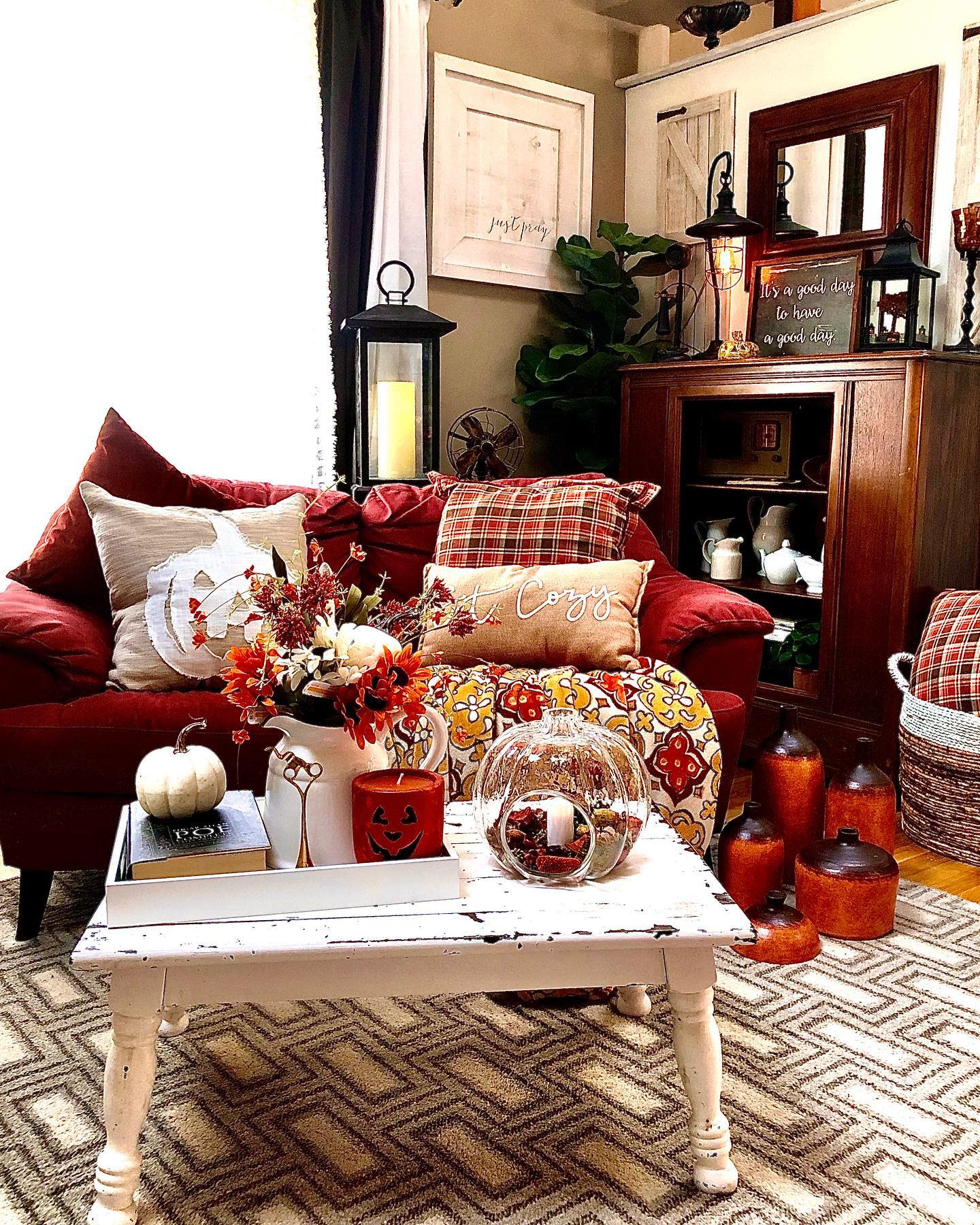 Cozy living room with a red sofa, autumn-themed decorations, pillows, lanterns, rustic cabinet, and a patterned rug