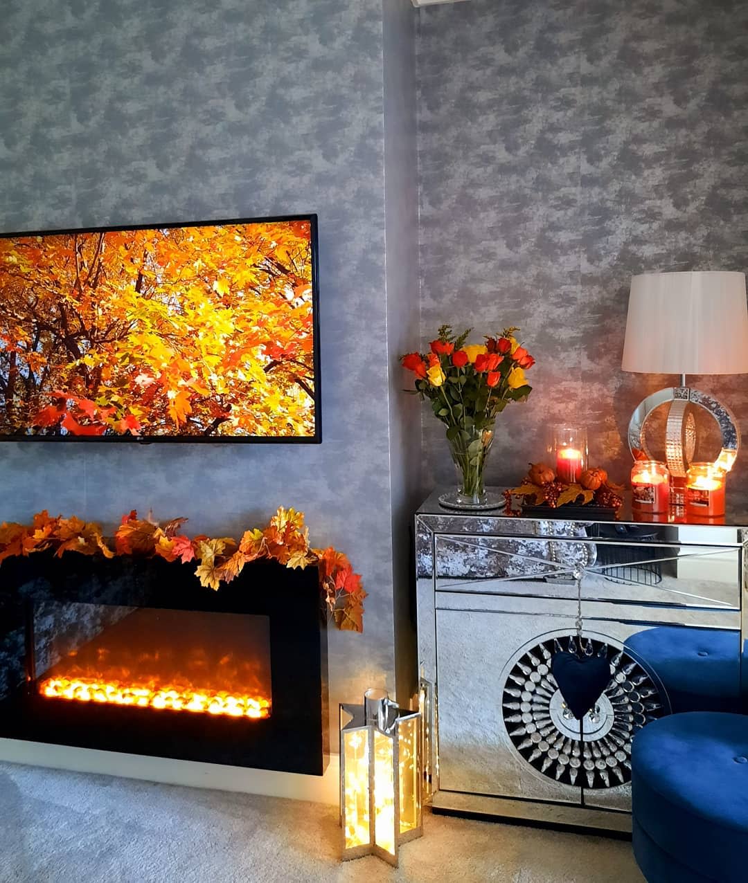 Cozy living room with autumn decor, featuring a lit fireplace, mirrored cabinet, fall leaves on TV, and orange flowers in a vase