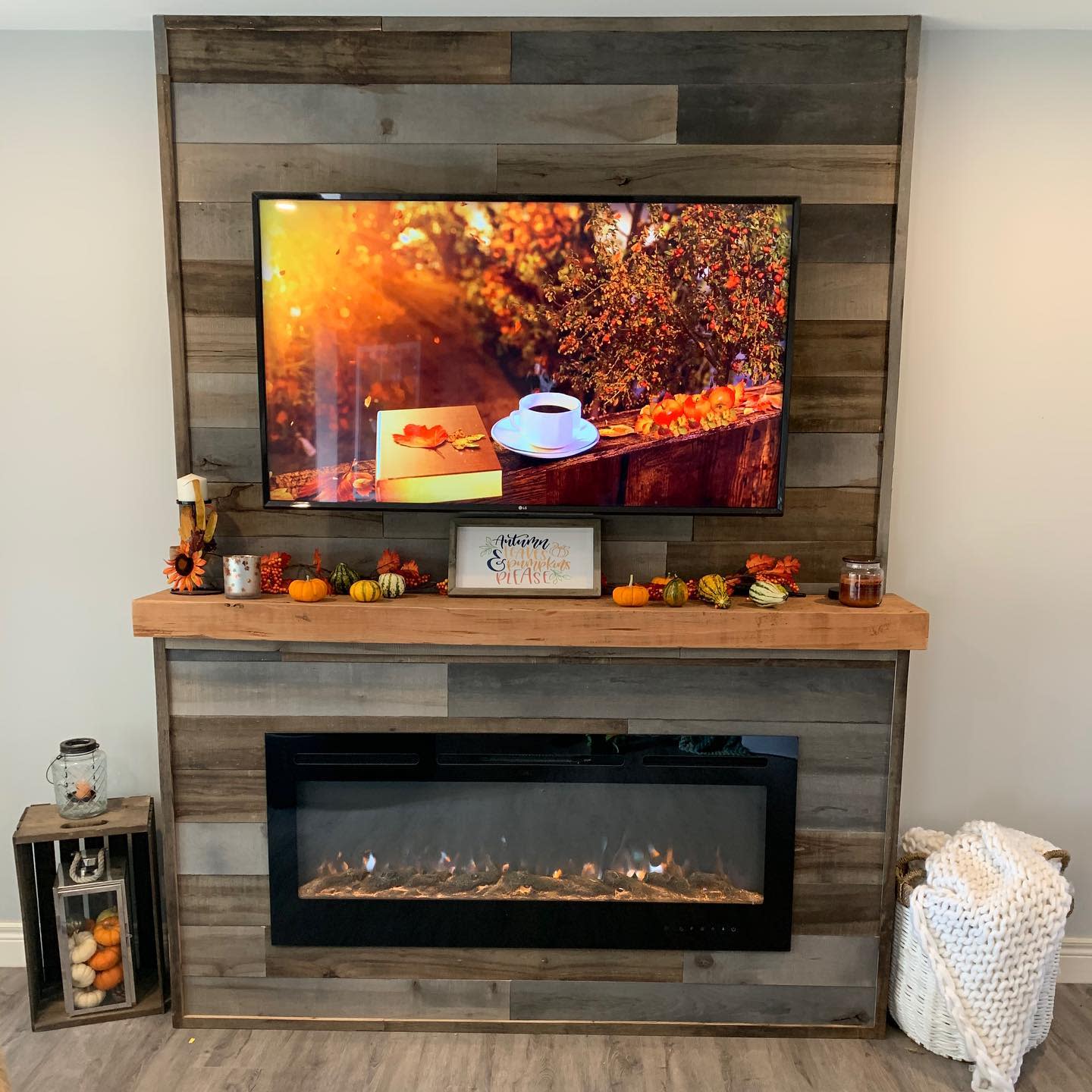 Wood-paneled wall with a mounted TV showing a fall scene, a shelf with autumn decor, and a modern electric fireplace below