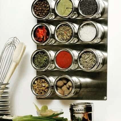 Mounted spice rack with clear-lid jars filled with various colorful spices and herbs, next to kitchen utensils on a counter