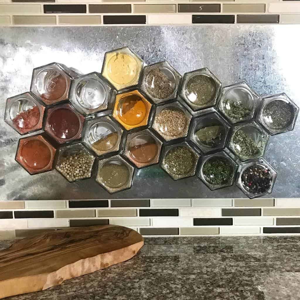 Hexagonal glass jars with various spices arranged on a metallic backsplash in a kitchen, above a granite countertop and wooden board