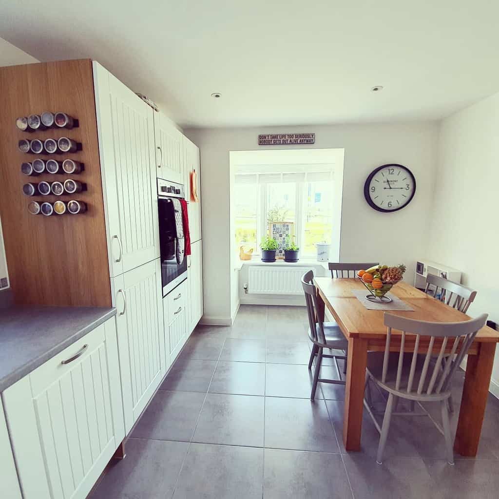 Bright kitchen with white cabinets, wall-mounted spice rack, wooden dining table, chairs, fruit bowl, and large wall clock