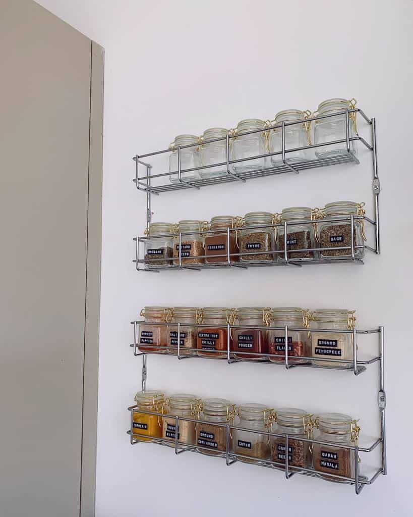 Three metal racks on a wall holding glass jars filled with various spices, each labeled with black tags