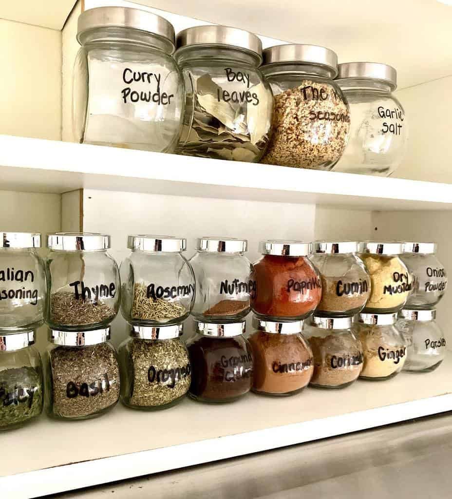 Labeled glass jars filled with various spices and herbs on white kitchen shelves