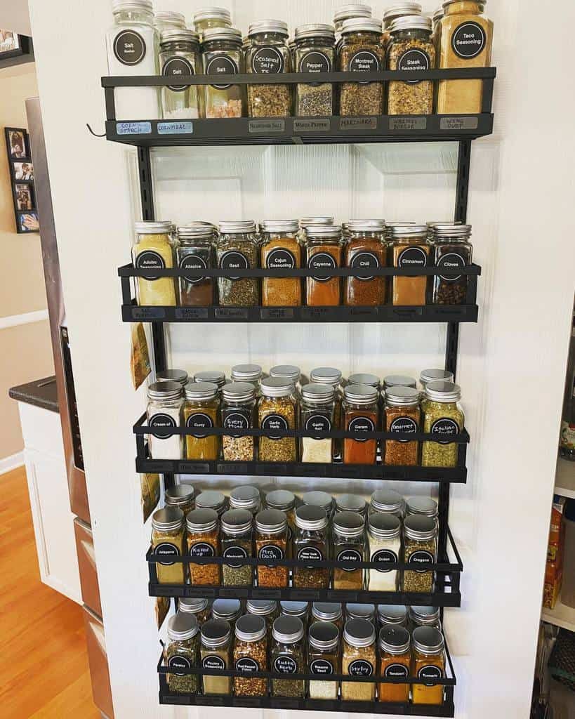 Wall-mounted spice rack with five shelves holding glass jars of various spices, each labeled with a black sticker