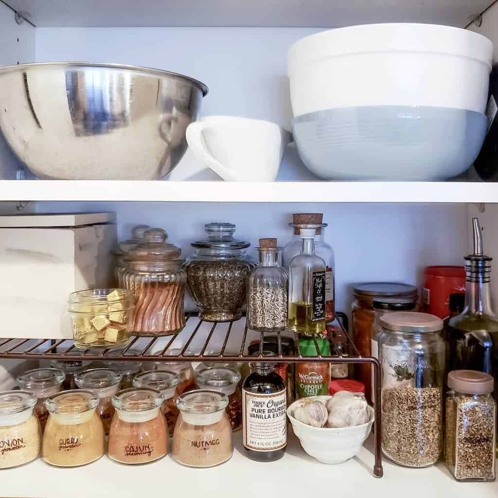 A well-organized pantry shelf with spices, herbs, glass jars, mixing bowls, and a mug, neatly arranged