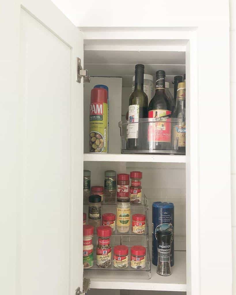 Open cupboard with three shelves holding olive oil spray, wine bottles, and various spice jars