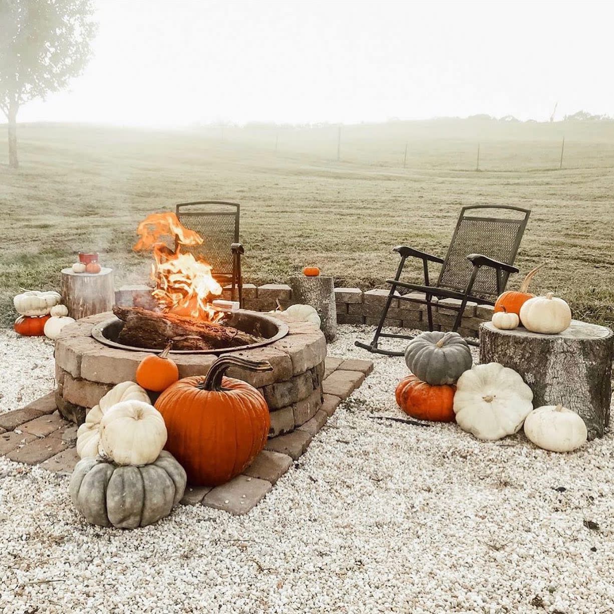 Outdoor fire pit with pumpkins, chairs, and logs on a gravel ground in a grassy field