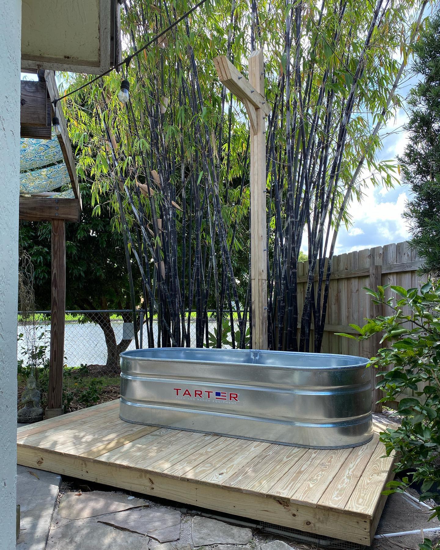 Galvanized metal tub on a wooden deck surrounded by bamboo and a fence, under a clear sky