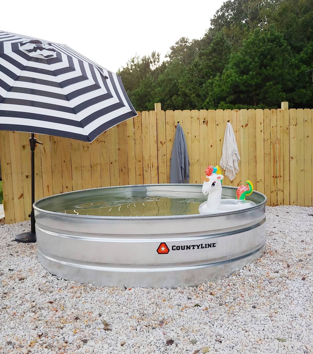 Galvanized stock tank pool with a striped umbrella, unicorn float, and towels on a fence; pebbled ground, trees in the background