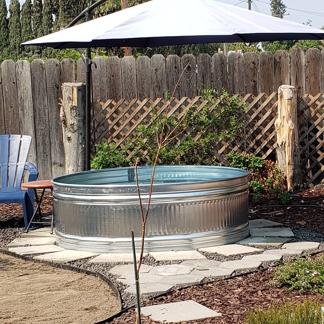 Galvanized metal stock tank pool under a white umbrella in a backyard, surrounded by wooden fences and a blue chair