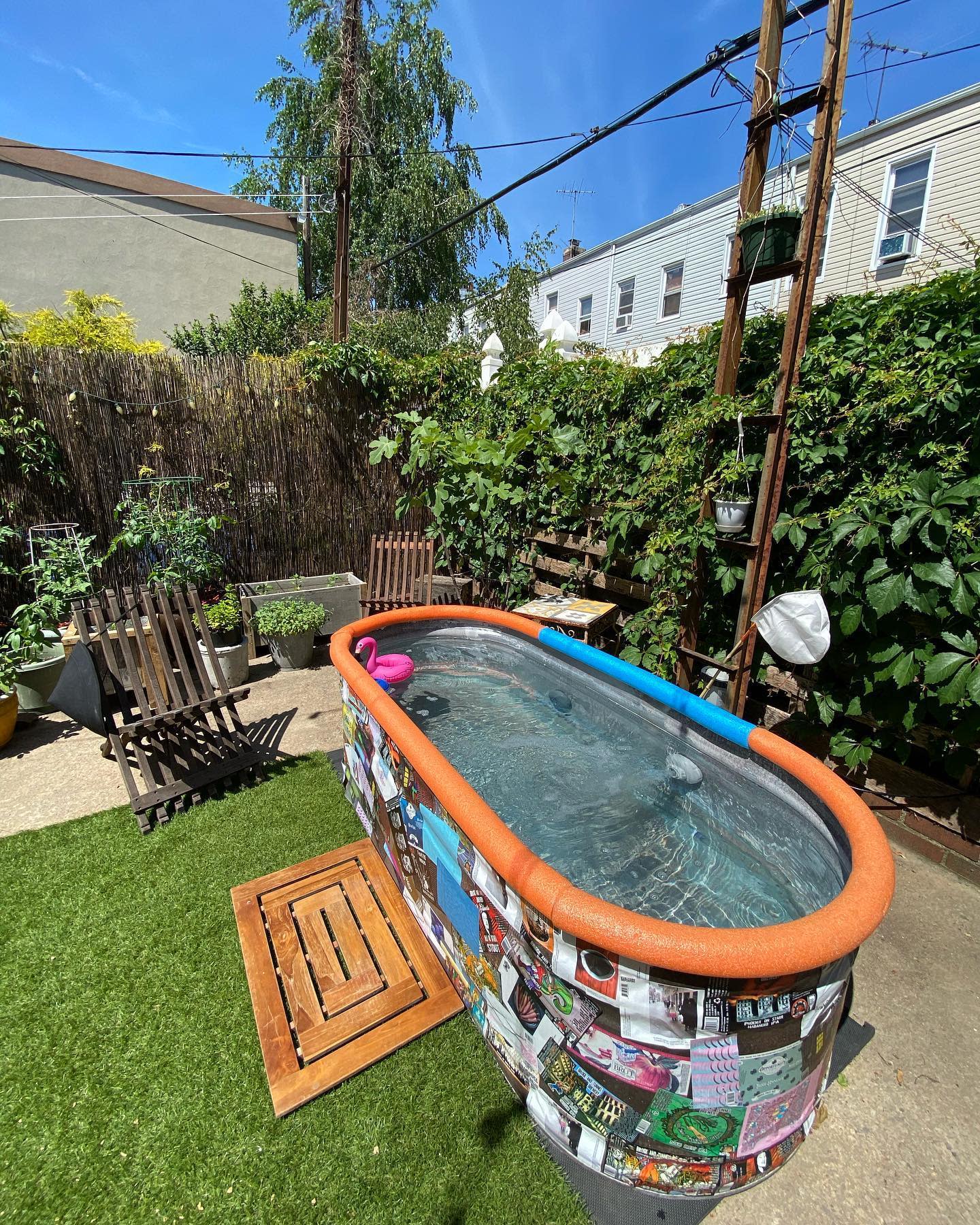 Eclectic backyard with an oval stock tank pool decorated with magazine clippings, foam pool noodles, wooden chairs, and lush greenery