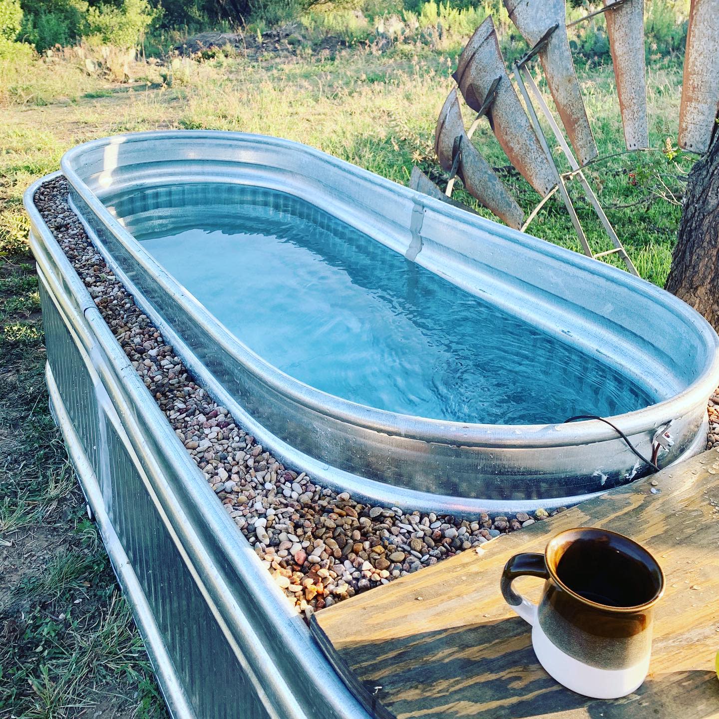 Galvanized metal trough filled with water next to a windmill, surrounded by pebbles