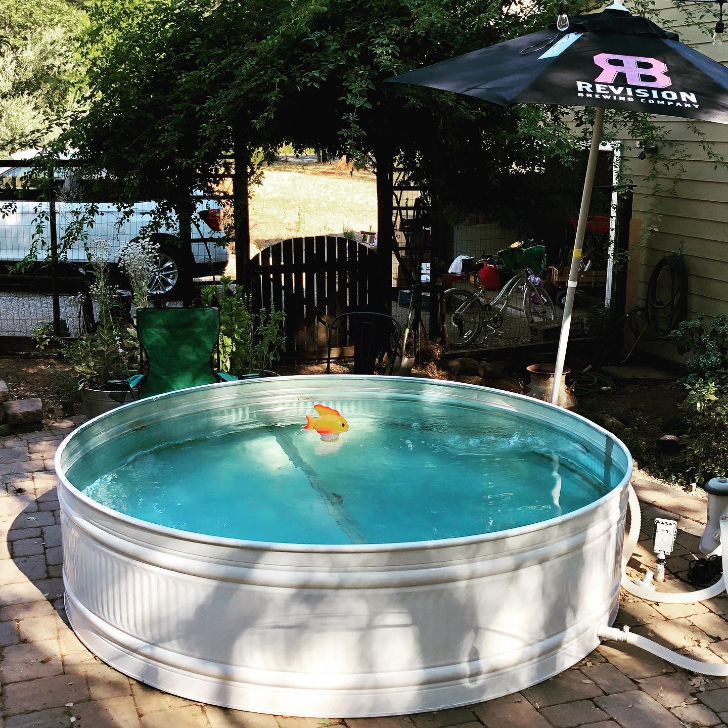 Simple backyard stock tank pool with a white exterior, brick patio, umbrella for shade, and a floating fish toy for a relaxed outdoor setup