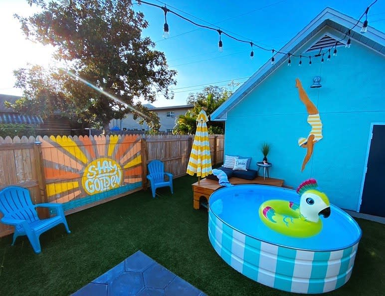 Colorful backyard with a small pool, inflatable parrot, beach chairs, striped umbrella, "Stay Golden" mural, and string lights