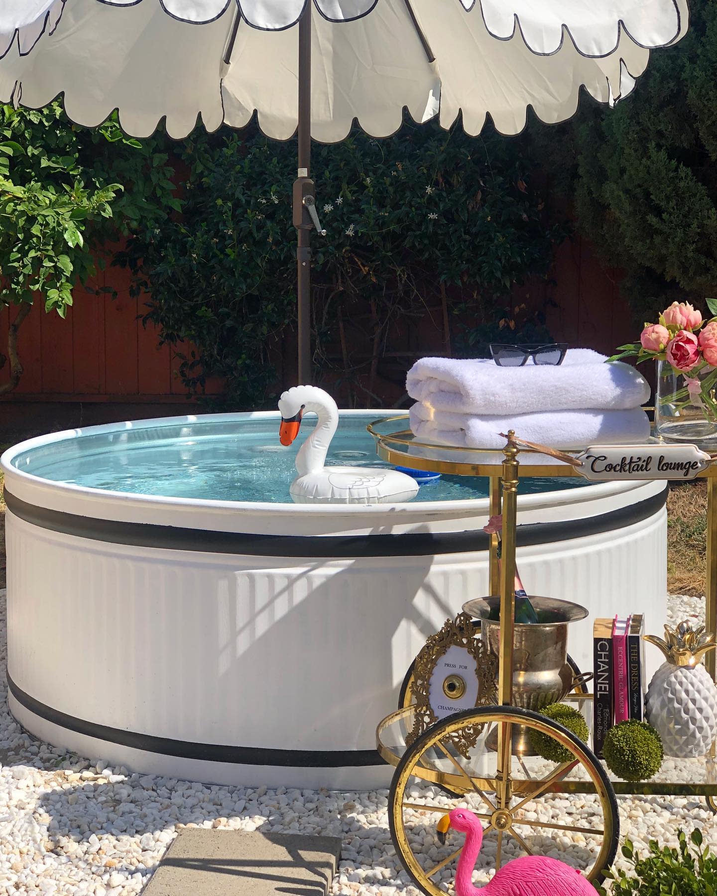 Backyard scene with a small pool, inflatable swan, umbrella, and a cart holding towels and flowers, with a pink flamingo decoration nearby