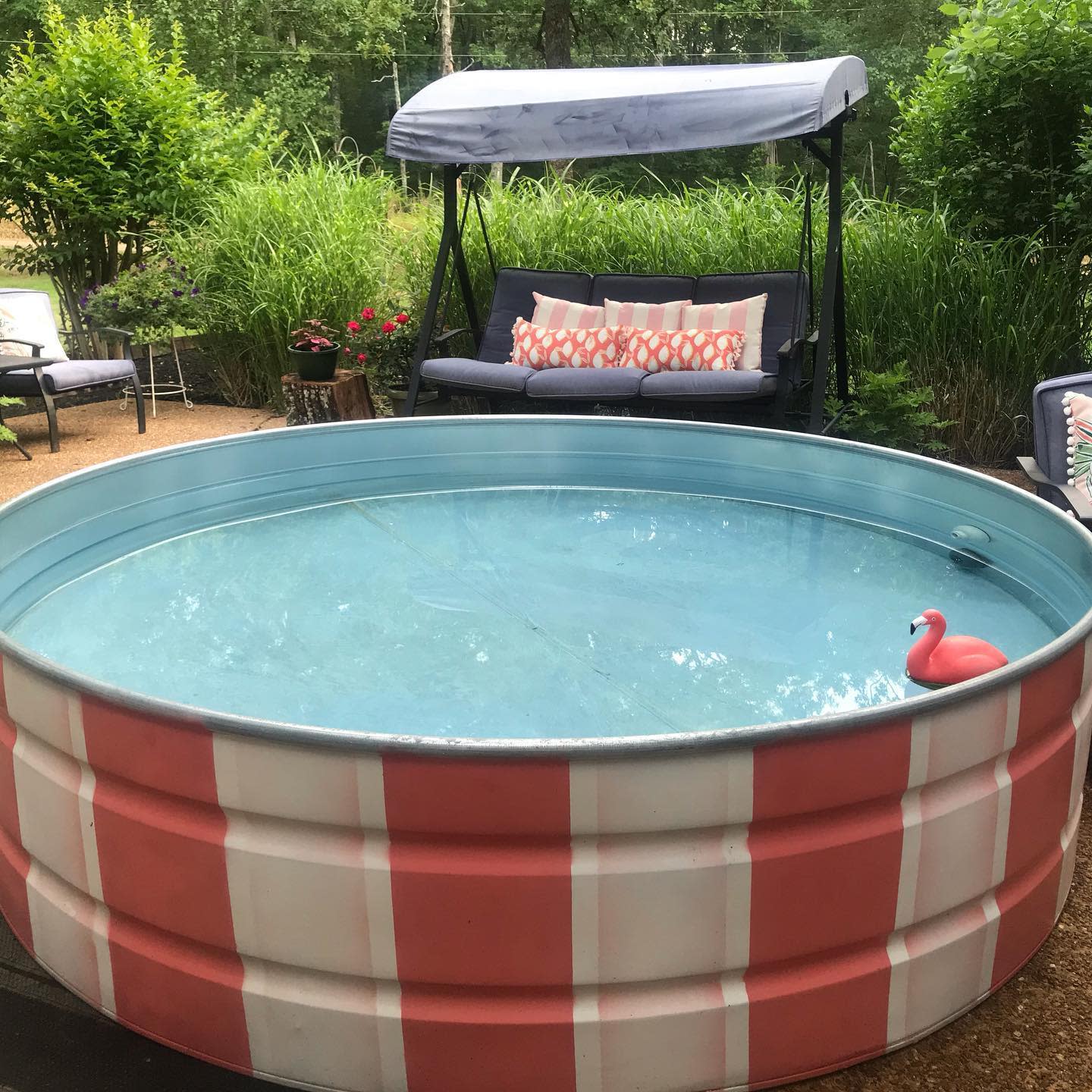 Round pool with a pink stripe and a small inflatable flamingo Nearby, a swing with pillows and green plants is in the background