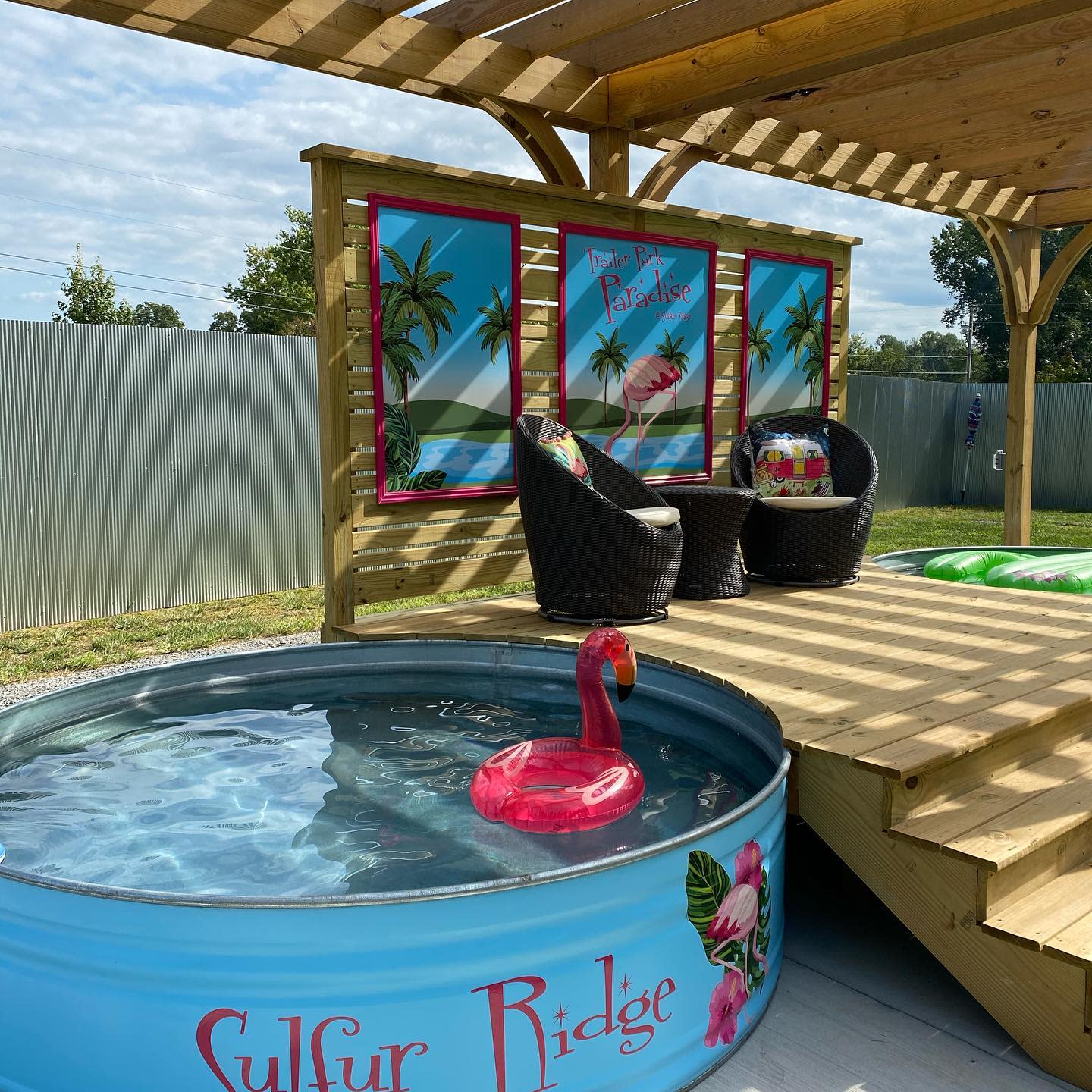 Tropical-themed backyard with a painted stock tank pool, wooden pergola, wicker seating, flamingo decor, and colorful wall art