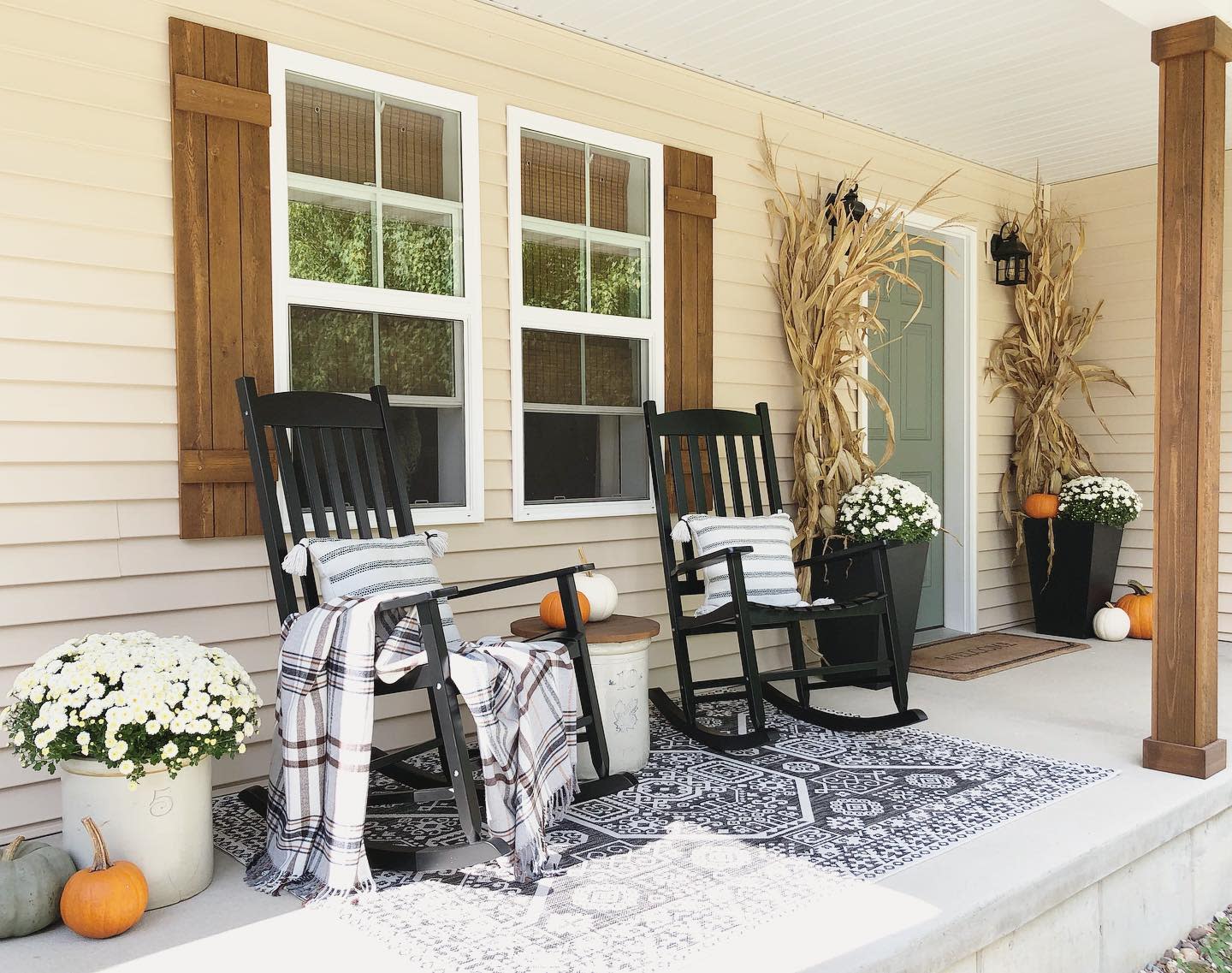 Front porch with two black rocking chairs, plaid blankets, fall decor, corn stalks, white mums, pumpkins, and patterned rug