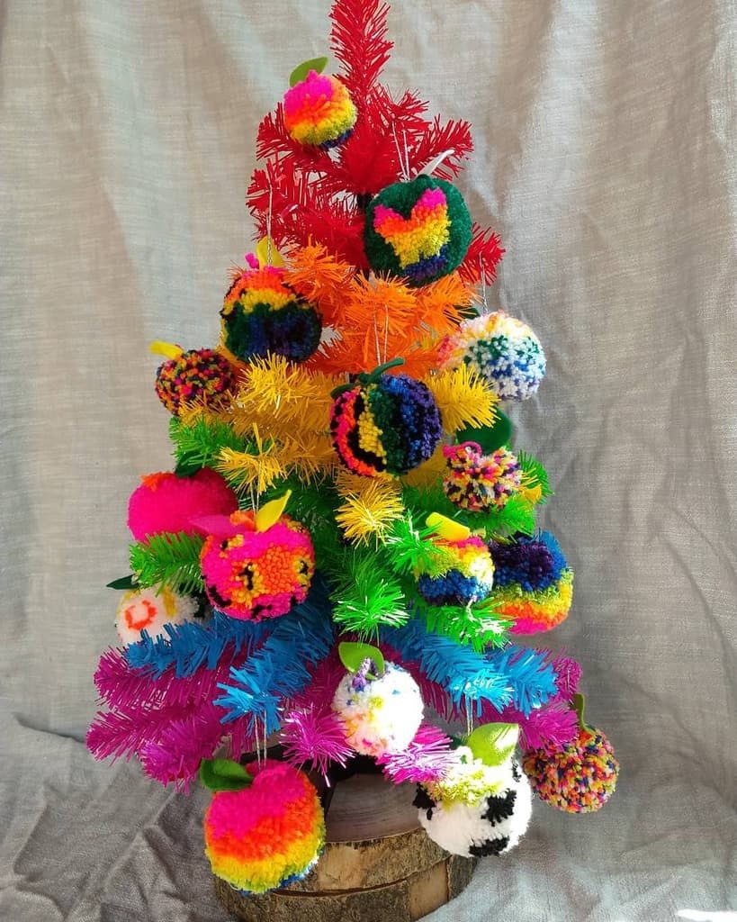 A small, colorful Christmas tree with multicolored pom-pom ornaments on a wooden base against a gray fabric background