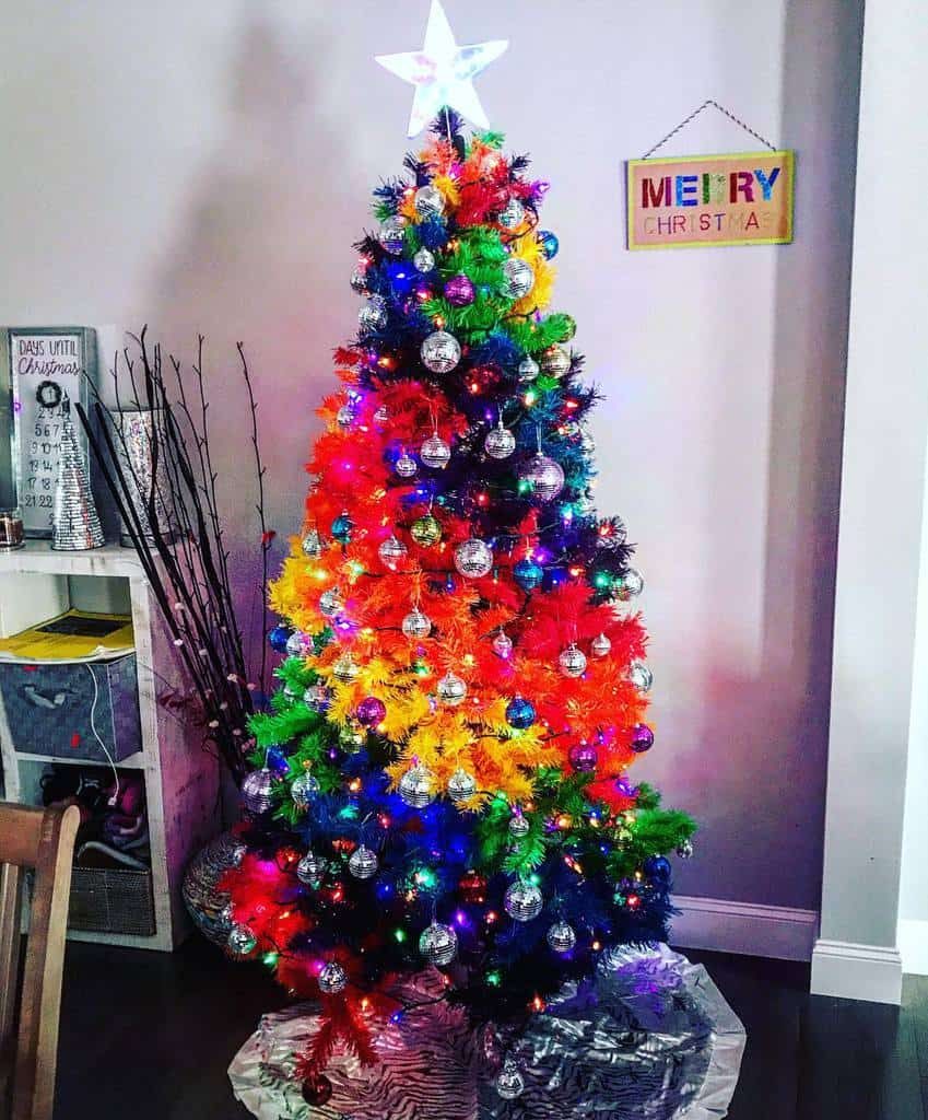 A rainbow Christmas tree decorated with silver baubles and colorful lights stands in a room with a "Merry Christmas" sign