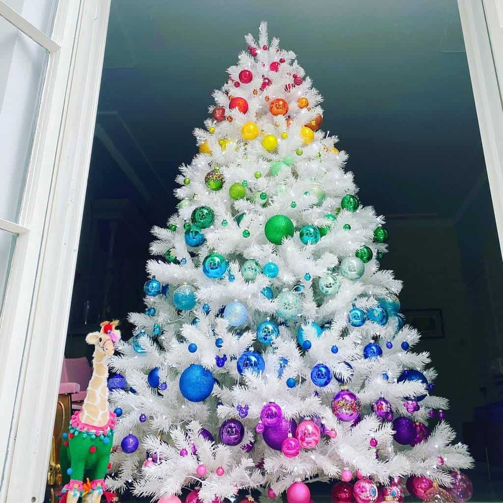 White Christmas tree decorated with rainbow-colored ornaments, with a small giraffe plush toy beside it