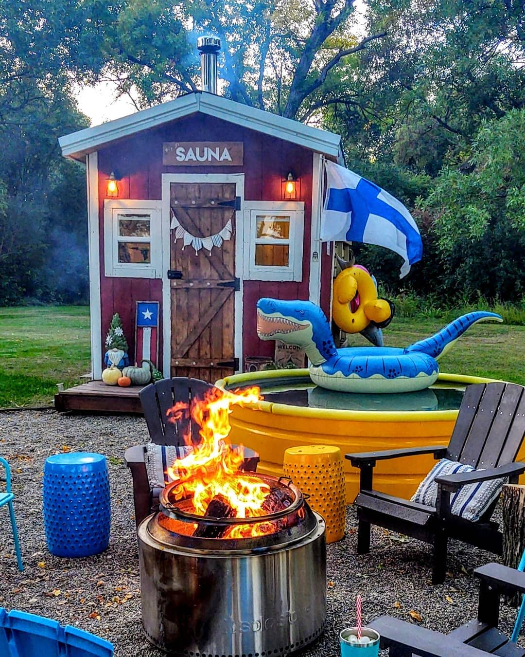 Rustic backyard with a red sauna cabin, Finnish flag, yellow stock tank pool, fire pit, Adirondack chairs, and inflatable pool floats