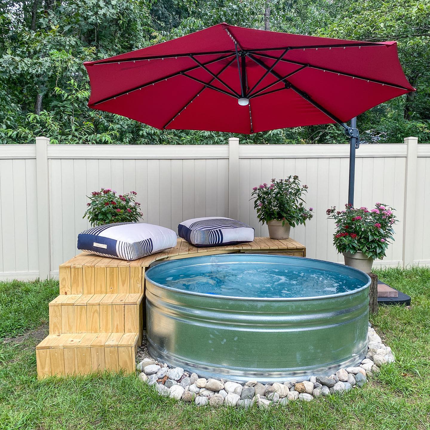 A small round pool with wooden steps, two striped cushions, a red umbrella, and potted plants in a fenced yard