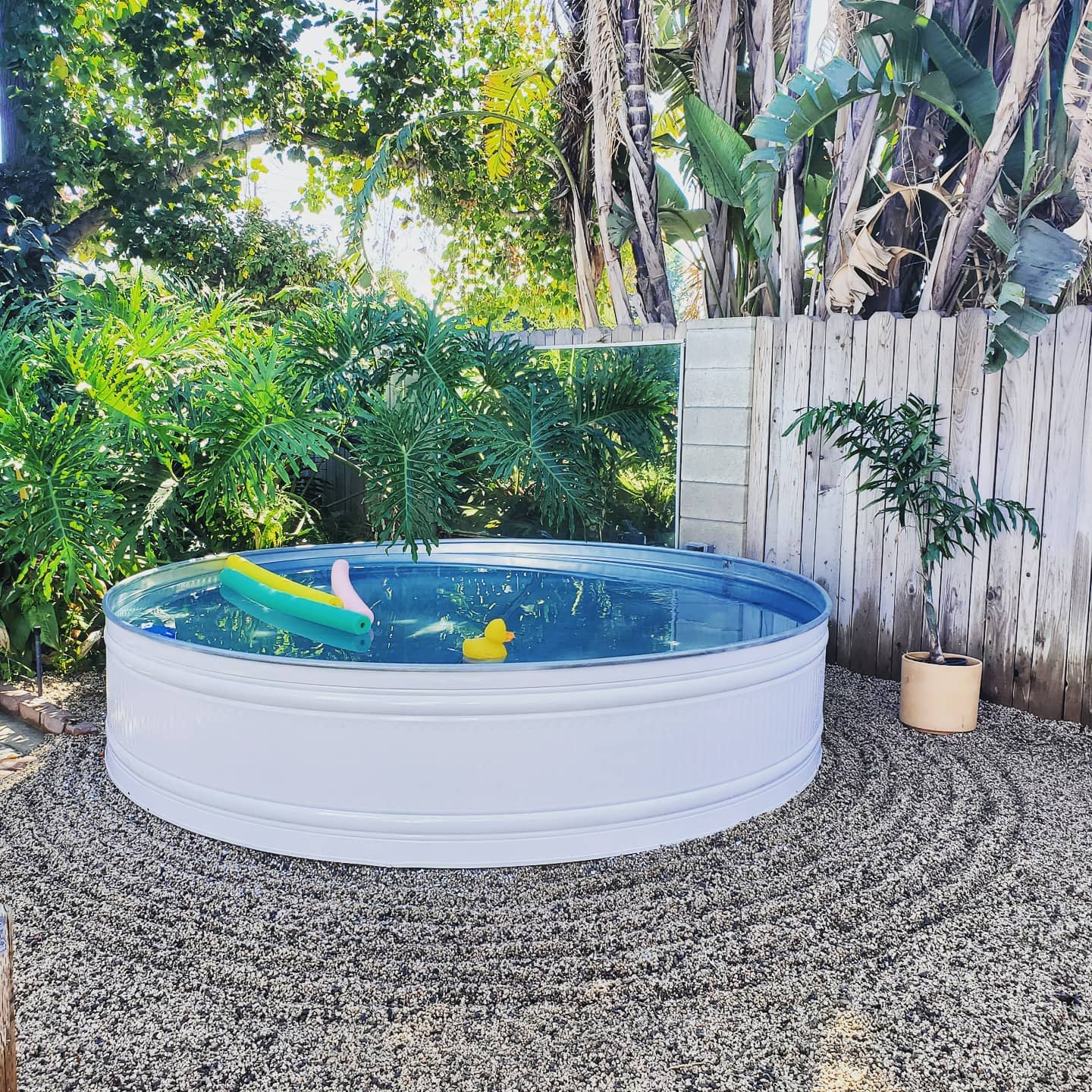 A small round pool with inflatable toys is surrounded by greenery and a wooden fence, set on a textured stone ground pattern