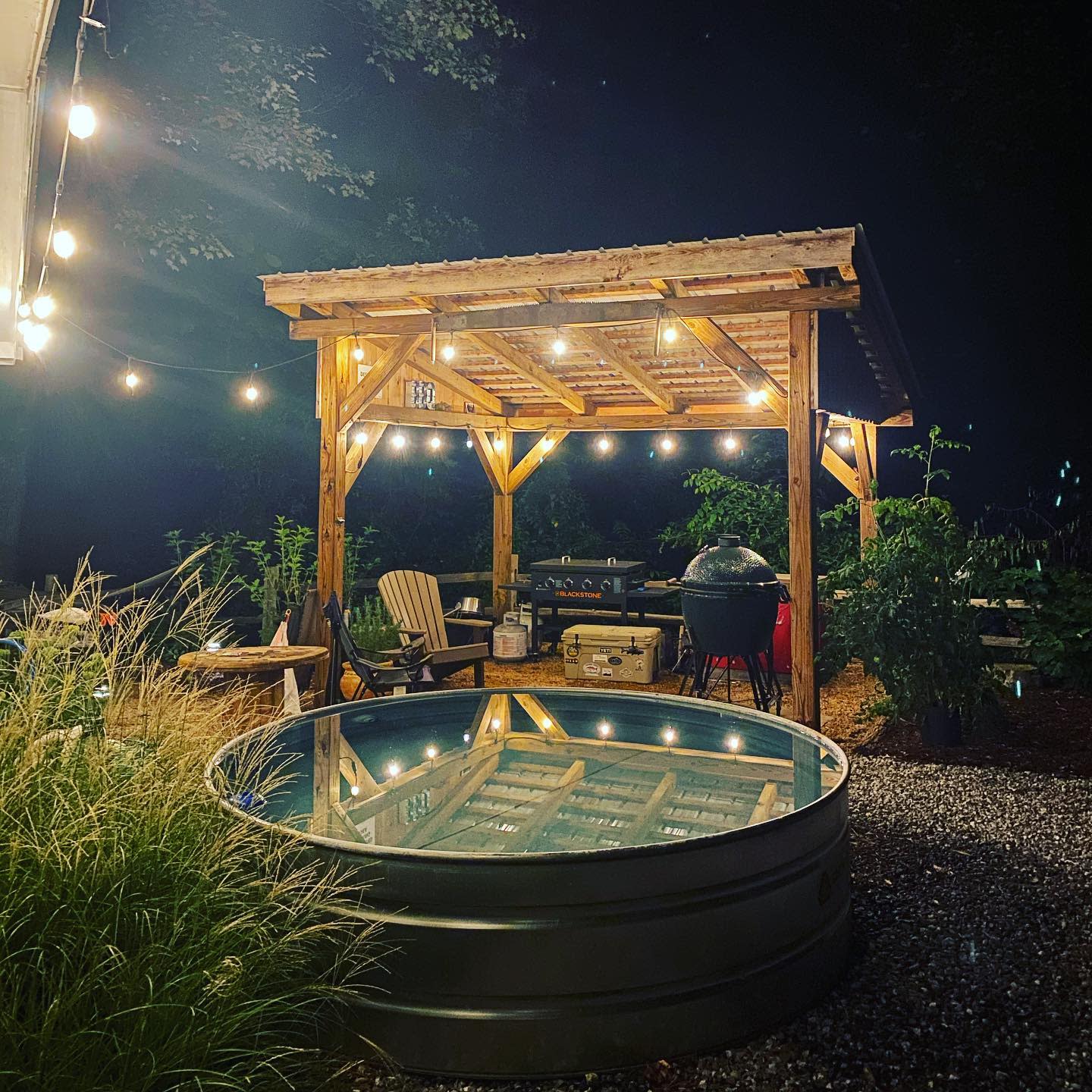 Cozy backyard with a stock tank pool, wooden pergola, string lights, and a grilling area, creating a warm ambiance at night